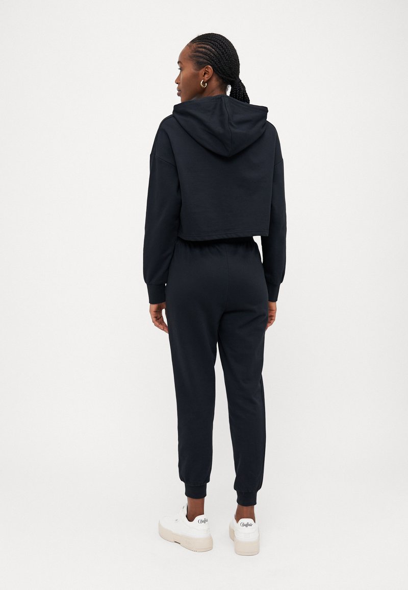 Felpa nera corta con cappuccio, abbinata a pantaloni della tuta neri coordinati. Realizzata in tessuto morbido, presenta polsini a costine e una vita elastica. Sneakers bianche.