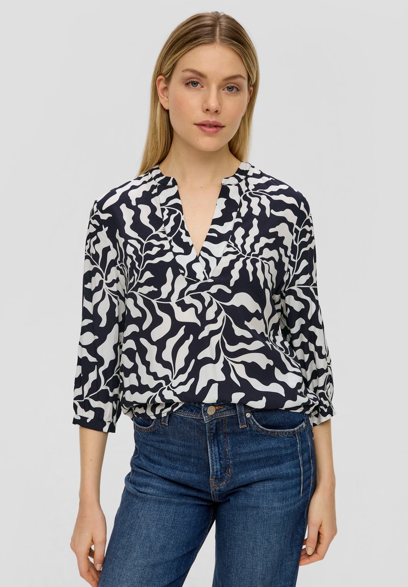 Blouse bleu marine avec des motifs abstraits blancs, décolleté en V et manches 3/4, fabriquée en tissu lisse ; associée à un jean en denim bleu.