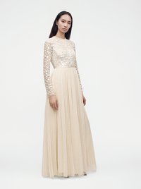 Femme portant une longue robe beige avec des manches transparentes à motifs et une jupe fluide, debout devant un fond blanc uni.