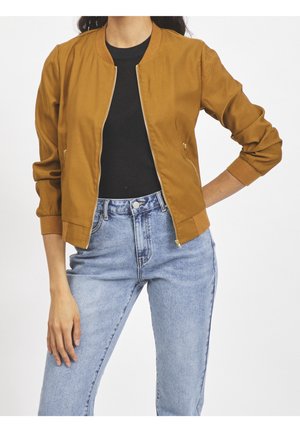 Frau trägt eine senffarbene Bomberjacke, ein schwarzes Top und hellblaue Jeans mit hoher Taille, eine Hand in der Hüfte, die andere entspannt.