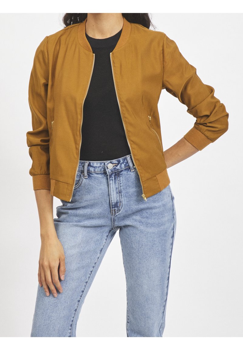 Femme portant une veste bomber moutarde, un haut noir et un jean taille haute bleu clair, une main sur la hanche et l'autre détendue.