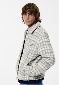 Harmaa-valkoinen ruudullinen takki pehmeällä tekstuurilla, jossa on terävä kaulus, vetoketjullinen edusta ja kaksi sivutaskua. Rentoa istuvuutta ja näkyvät saumat.