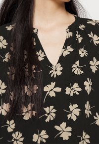 Blouse noire avec un décolleté en V, ornée d'un motif floral beige clair. Le tissu semble lisse avec un léger éclat. Manches courtes froncées.