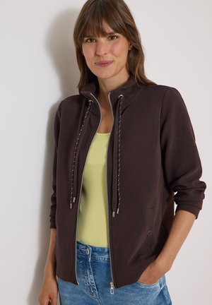 Kvinde iført en mørkebrun lynlåstrøje over en gul top og blå jeans, stående med den ene hånd i lommen mod en lys baggrund.