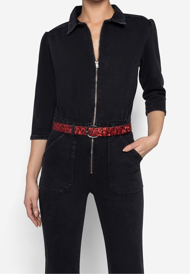 Femme portant une combinaison en jean noire avec manches trois-quarts et fermeture éclair frontale, accessoirisée d'une ceinture rouge à motifs.