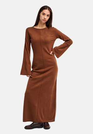 Femme portant une longue robe marron avec des manches évasées et des chaussures noires, debout avec une main sur la hanche devant un fond uni.