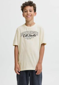 Kremskrāsas T-krekls ar īsām piedurknēm, ar melnu teksta dizainu: "JACK&JONES Coll. Studio" ar papildu detaļām zem tā.