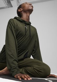 Dunkelgrüner Sporthoodie mit schwarzen Kordeln, ausgestattet mit einem strukturierten Stoff und einem Logo. Kombiniert mit passenden lässig geschnittenen Hosen auf einer Yogamatte.