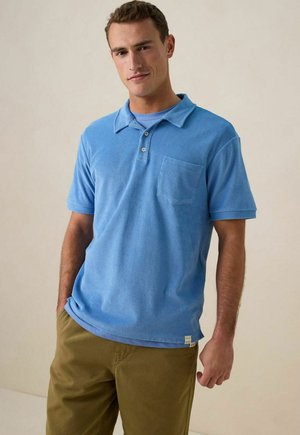 Lichtblauwe terry polo shirt met een kraag, korte mouwen en een zak op de borst. Zachte textuur en relaxte pasvorm. Gepaard met kaki broeken.
