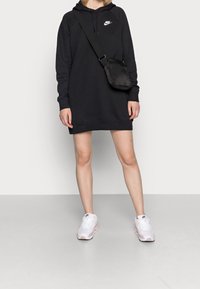 Vestido sudadera negro con bolsillo frontal y capucha, que presenta un logo de Nike en blanco. Combinado con zapatillas Nike blancas y rosas y un bolso bandolera negro.