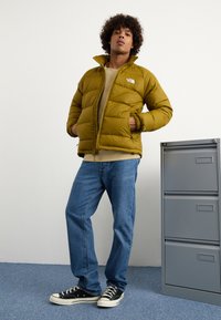 Puffervest i olivengrøn med lynlås fortil, har en høj krave og sidelommer. Båret over en beige skjorte, parret med blå jeans og sorte sneakers.