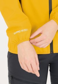 Gul vandtæt jakke med elastiske manchetter, udstyret med "W-PRO 15000" i hvid tekst. Glat tekstur, frontlynlås, kontrasteret med sorte bukser.