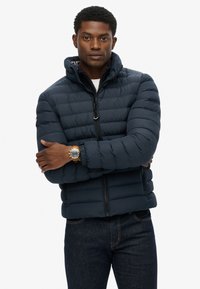 Mørkeblå polstret jakke med lynlås fortil, justerbar hætte og elastiske manchetter. Kombineret med mørkeblå jeans og et guld ur.