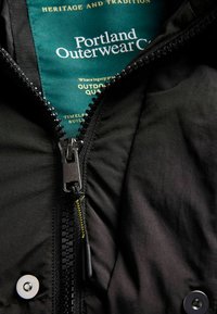 Černá vnější bunda s aplikovaným zipem se černými zuby, zelenou podšívkou s nápisem "Portland Outerwear Co" a stříbrnými knoflíky.