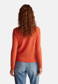 Kobieta z długimi kręconymi rudymi włosami, ubrana w pomarańczowy sweter z długim rękawem i niebieskie dżinsy, widziana od tyłu.