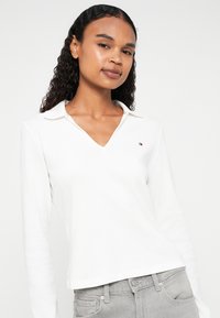 Maglietta bianca a maniche lunghe con scollatura a V, dal taglio rilassato e con colletto, dotata di un piccolo logo ricamato sul lato sinistro del petto.