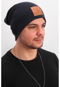 Navy knitted beanie with a slouchy fit, featuring a rectangular brown leather patch with logo detail. Model wears a black textured shirt.