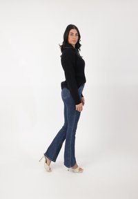 Suéter negro de punto, jeans azul de campana y sandalias de tacón blanco. Las texturas suaves y el diseño ajustado destacan el estilo casual del atuendo.