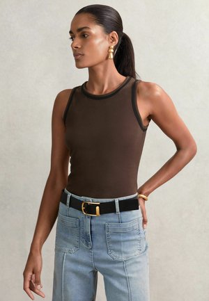 Kvinde med mørk hestehale iført brun ærmeløs top, lyseblå jeans med store lommer, sort bælte med guldfarvet spænde og guldringe.
