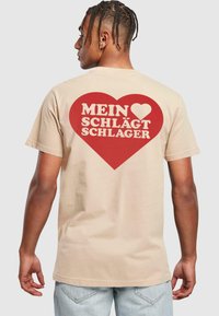 Šviesiai rudos spalvos medvilnės marškinėliai su dideliu raudonu širdies piešiniu ant nugaros, kuriame balta tekstas skaitomas "MEIN ♥ SCHLÄGT SCHLAGER." Trumpomis rankovėmis ir laisvu siluetu.