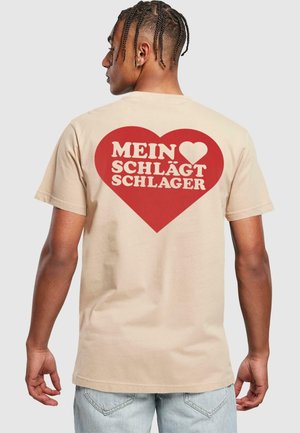 Beige t-paita, jossa on selässä suuri punainen sydängrafiikka ja valkoinen teksti "MEIN ♥ SCHLÄGT SCHLAGER." Lyhyet hihat ja rento malli.