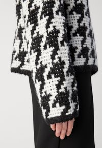 Black and white patterned sweater with textured knit, featuring a loose fit and ribbed cuffs. Sleeve extends past the wrist.