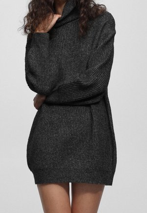 Femme portant une robe pull en maille côtelée gris foncé à manches longues, debout devant un fond clair uni.