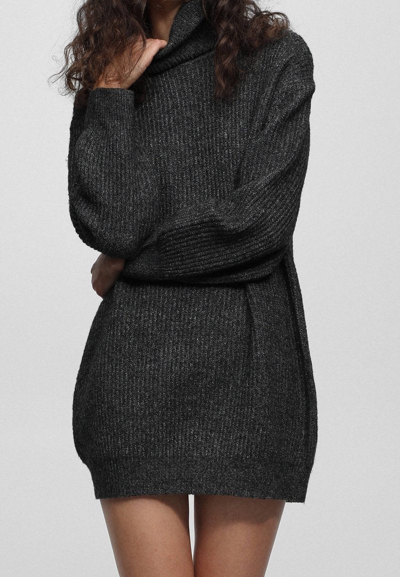 Femme portant une robe pull en maille côtelée gris foncé à manches longues, debout devant un fond clair uni.
