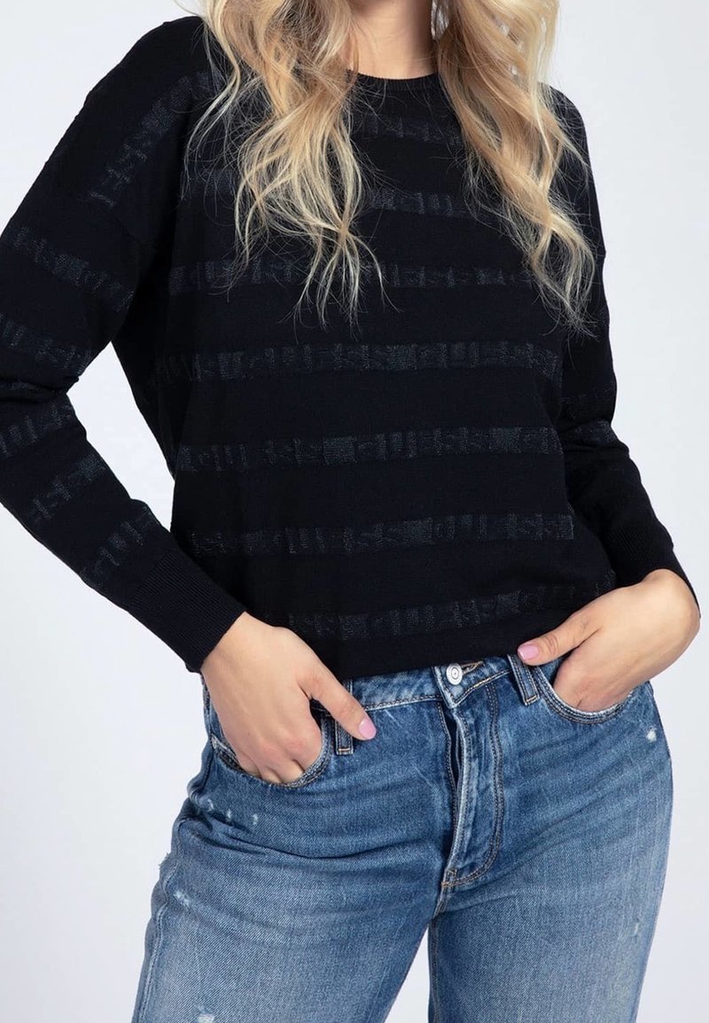 Femme aux longs cheveux blonds portant un pull rayé noir et un jean bleu, mains dans les poches, sur un fond clair.