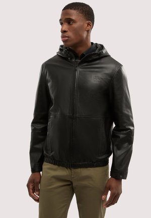 Hombre con chaqueta negra de cuero con cremallera y capucha, y pantalones caqui, mirando hacia la izquierda sobre un fondo liso.