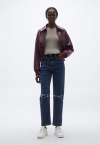 Bordó bőrkabát kivágva egy texturált krémszínű kötött pulóver fölött, kék szakadt farmerrel és fehér sportcipővel párosítva.