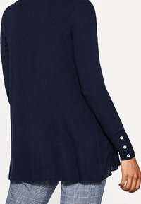 Marineblaues Langarmoberteil mit lockerer Passform, rundem Halsausschnitt und Knopfdetails an den Ärmeln. Weicher Stoff, leichtes Material und seitliche Schlitze.