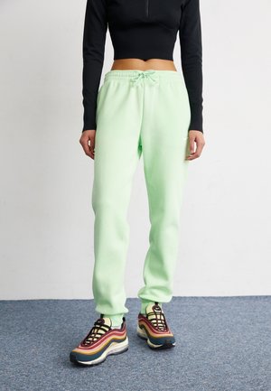 Personne portant un pantalon de jogging vert pastel, des baskets multicolores et un haut court noir à manches longues, debout sur un tapis bleu contre un mur blanc.