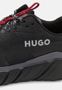 Baskets de sport noires avec accents gris, tige en tissu texturé, logo réfléchissant "HUGO" et lacets rouges et gris. Semelle au design profilé.