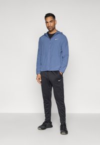 Αναμνηστική ελαφριά μπλε ζακέτα Nike με κουκούλα, μαύρο αθλητικό παντελόνι και μαύρα sneakers. Διαθέτει το λευκό λογότυπο της Nike στη ζακέτα και στο παντελόνι.