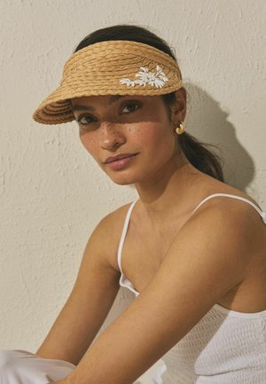 Frau mit dunklen Haaren, die ein weißes Tanktop trägt und eine beige gewebte Schirmmütze mit weißer Blumenstickerei aufhat, sitzt an eine beige strukturierte Wand gelehnt.