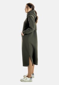 Largo vestido sudadera verde oliva con capucha, con aberturas laterales y bolsillos, hecho de una tela suave. Combinado con zapatillas blancas con detalles en azul.