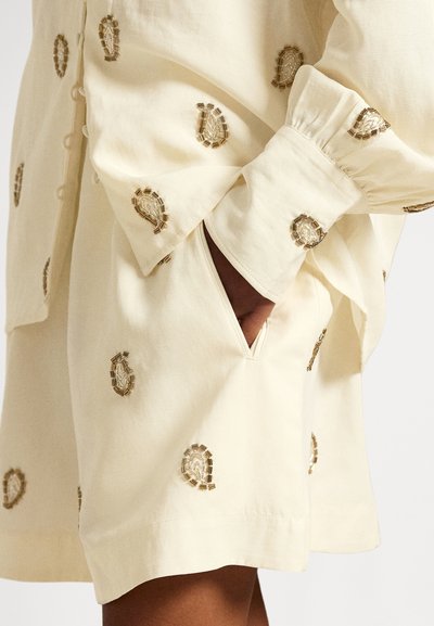 Šviesiai geltona marškinėliai su siuvinėtais dekoratyviniais elementais, laisvo silueto, su sagų užsegimu ir šonine kišenės detale.