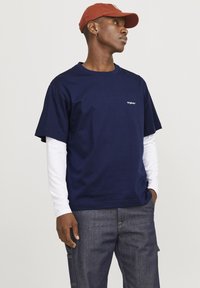 Mørkeblå bomull T-skjorte lagd over en hvit longsleeve skjorte, med en liten logo på brystet og en avslappet passform. Brun caps brukt.