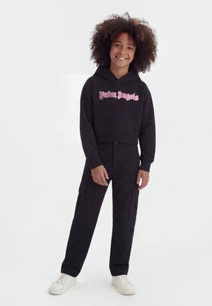 Copil cu părul creț, purtând un hanorac scurt negru cu text roz, pantaloni cargo negri și adidași albi, zâmbind și stând în picioare.