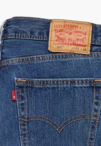 Blauwe spijkerbroek van denim met een tan leren label waarop rode tekst staat. De stiksels zijn in een lichtere contrasterende kleur en er is een klein rood label zichtbaar aan de achterkant.
