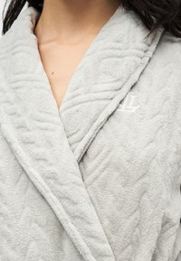Peignoir gris clair doux avec motif texturé et lettre L brodée sur la poitrine, porté par une personne à la peau brune et aux cheveux foncés.