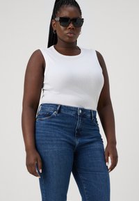 Top blanco de canalé combinado con jeans de tiro alto azules oscuros. Las gafas de sol negras y el cabello trenzado añaden contraste al atuendo.