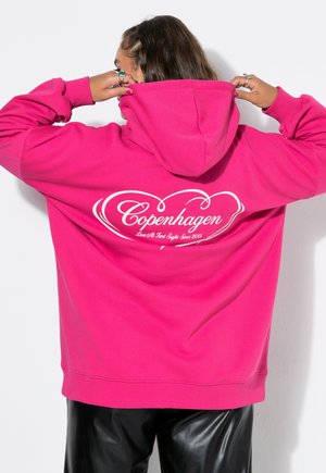 Person justerer hætten på en lys pink hættetrøje med hvid tekst, der lyder "Copenhagen Love At First Sight Since 2015" på ryggen.