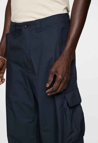 Marineblaue Cargo-Hose aus strukturiertem Stoff, mit Knopfverschluss und Seitentasche, gezeigt von der Taille bis zur Mitte des Oberschenkels.