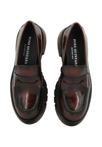Mocasines de cuero marrón con un acabado brillante, que cuentan con una superficie texturizada, un detalle decorativo de lazo en la parte delantera y una suela negra de goma gruesa.