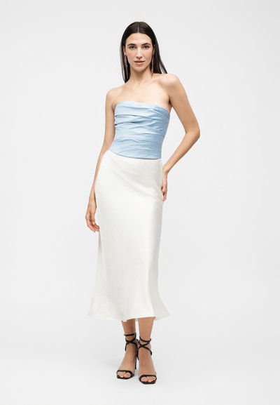 Γυναίκα που φοράει έναν αμάνικο γαλάζιο ρούχο με πιέτες, μακριά λευκή φούστα και μαύρα λουστρίνι τακούνια με κορδόνια, στέκεται με το ένα χέρι στη μέση.