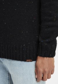 Sort strikket sweater med multicolor pletter. Har ribbede manchetter og kant. Lille brun label nederst til venstre. Båret over lys denim.