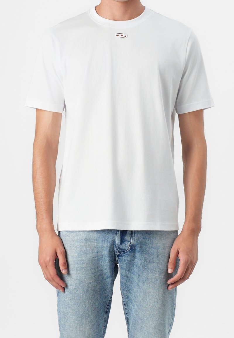 Homme portant un t-shirt blanc uni à manches courtes et un jean bleu clair, debout devant un fond blanc.