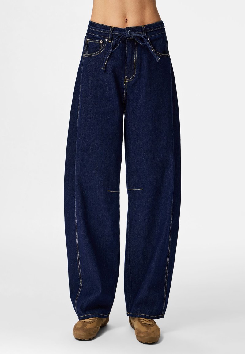 Pantalones de mezclilla de pierna ancha en azul oscuro con costuras amarillas contrastantes. Cuentan con cintura con cordón y bolsillos funcionales en la parte delantera.